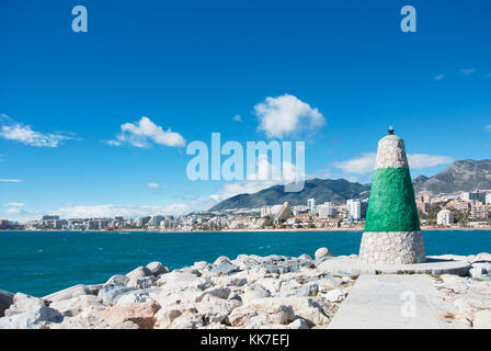 A little lighthouse of white and green color at the pier covered with stones and bright blue water, beach and hotels at the background. Copy space for Stock Photo