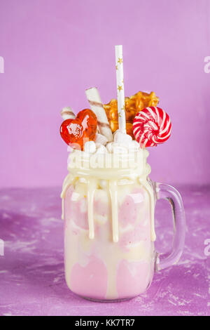 Freakshake from pink smoothie, cream. Monstershake with lollipops, waffles and marshmallow. Extreme milkshake in a Mason jar. Pink purple background Stock Photo