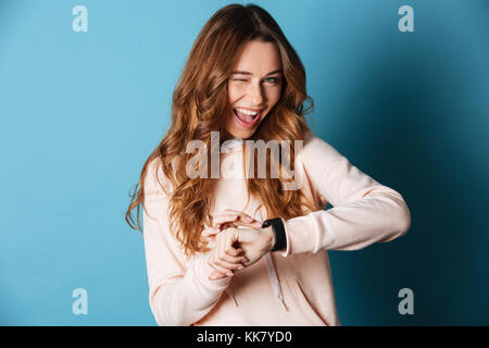 Image of happy young woman standing isolated over blue wall background. Looking camera with watch on hand winking. Stock Photo