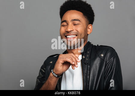 Portrait of a cheerful young african man in leather jacket laughing with eyes closed isolated over gray background Stock Photo