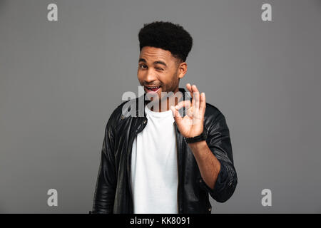 Young friendly african guy winks one eye while showing OK gesture, isolated on gray background Stock Photo