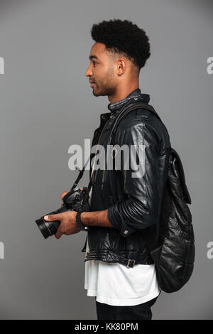 Side view photo of handsome stylish african man with backpack holding photo camera, isolated on gray background Stock Photo