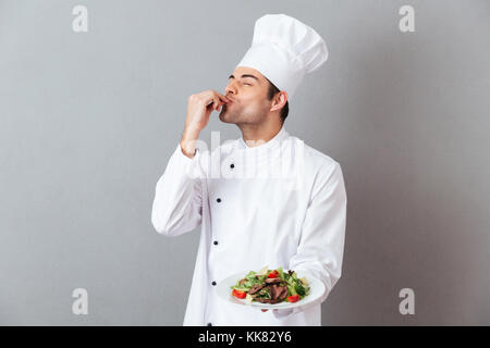 Portrait of a handsome male chef dressed in uniform holding plate with dish isolated over gray background Stock Photo