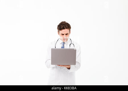 Portrait of a concentrated young male doctor with stethoscope dressed in uniform standing and using laptop isolated over white background Stock Photo