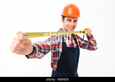 Portrait of an excited young male builder showing yellow measuring tape while standing and looking at camera isolated over white background Stock Photo