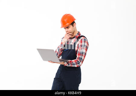 Portrait of a pensive young male builder working on laptop computer while standing isolated over white background Stock Photo