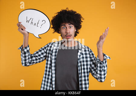 Portrait of a puzzled confused african man holding WHAT speech bubble and looking at camera isolated over orange background Stock Photo