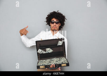 Portrait of a young afro american man in sunglasses pointing finger at briefcase full of money banknotes isolated over gray background