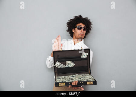 Portrait of a funny young afro american man in sunglasses holding briefcase full of money banknotes isolated over gray background