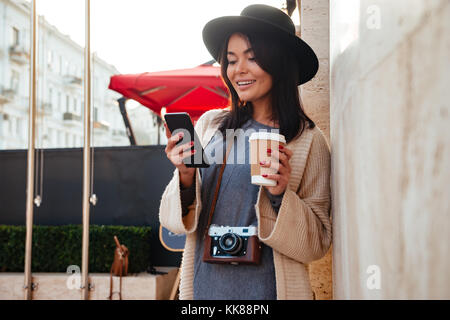 Young attractive asian woman with retro photocamera texting message and holding coffee cup while standing on the city street Stock Photo