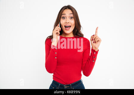 Portrait of a happy excited asian woman talking on mobile phone and having an idea while standing and pointing finger up isolated over white backgroun Stock Photo