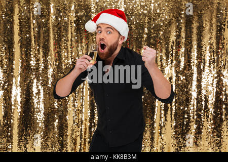 Portrait of a cheerful bearded man in christmas hat drinking champagne from a glass isolated over golden shiny bakground Stock Photo