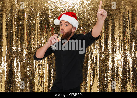 Portrait of a happy bearded man in christmas hat drinking champagne from a glass isolated over golden shiny bakground Stock Photo