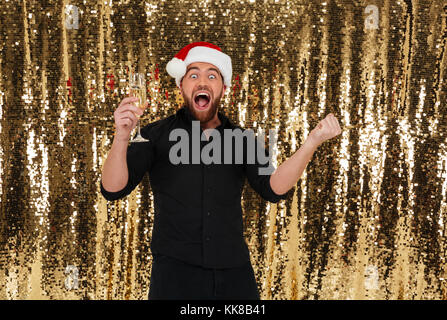 Portrait of an excited bearded man in christmas hat celebrating and drinking champagne from a glass isolated over golden shiny bakground Stock Photo