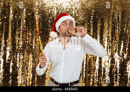 Portrait of a funny young man in christmas hat drinking champagne from a glass and holding a petard isolated over golden shiny bakground Stock Photo