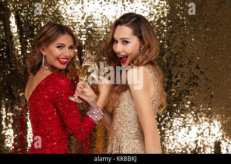 Portrait of two pretty smiling girls in shiny dresses holding glasses with champagne while toasting and celebrating isolated over golden shiny backgro Stock Photo