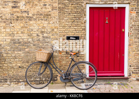Vintage bicycle with retro wicker basket parked in front of a house brick wall next to a bright red door Stock Photo