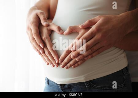 Close up portrait of man's and woman's hands making heart gesture over pregnant belly indoors Stock Photo