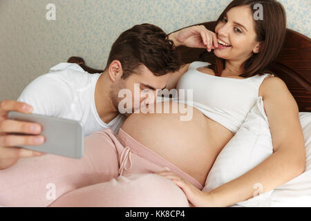 Lovely young pregnant couple taking selfie while lying in bed, man kissing pregnant belly Stock Photo