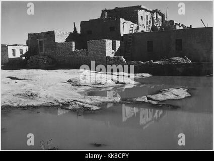 A black and white exterior photograph of an adobe house, the multistory home consists of several sections that are in various stages of completion, a reflection of the home can be seen in the pool of water in the foreground, the image comes from a series of photographs known as the mural project by Ansel Adams, he was commissioned by the National Park Service in 1941 to create a photographic mural for the Department of the Interior Building in Washington DC, the project came to an end due to World War II, the home is part of Acoma Pueblo and has been inhabited for over 800 years, Acoma Indian  Stock Photo