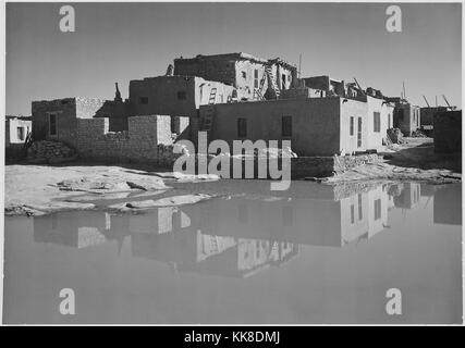 A black and white exterior photograph of an adobe house, the multistory home consists of several sections that are in various stages of completion, a reflection of the home can be seen in the pool of water in the foreground, the image comes from a series of photographs known as the mural project by Ansel Adams, he was commissioned by the National Park Service in 1941 to create a photographic mural for the Department of the Interior Building in Washington DC, the project came to an end due to World War II, the home is part of Acoma Pueblo which has been inhabited for over 800 years, Acoma India Stock Photo