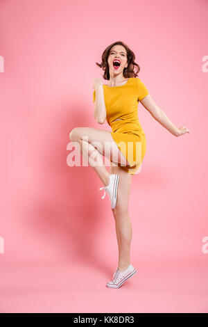 Full length portrait of pretty young woman in elegant yellow dress showing winner gesture, looking at camera, isolated on pink background Stock Photo