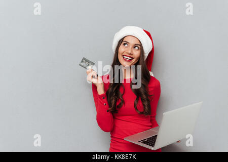 Charming brunette woman in Santa's hat and red blouse holding credit card and laptop computer, looking aside while chooses what to buy, isolated on gr Stock Photo