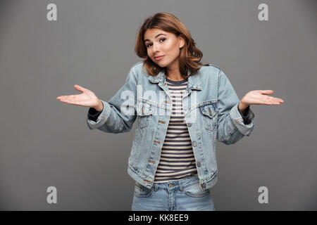 Portrait of a young doubtful teenage girl dressed in denim jacket shrugging shoulders and looking at camera isolated over gray background Stock Photo