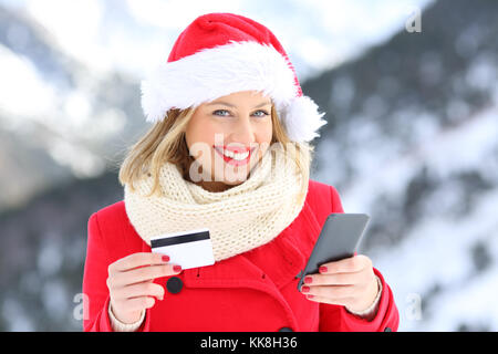 Happy girl on christmas wearing santa claus hat holding credit card and smart phone with a snowy mountain in the background Stock Photo
