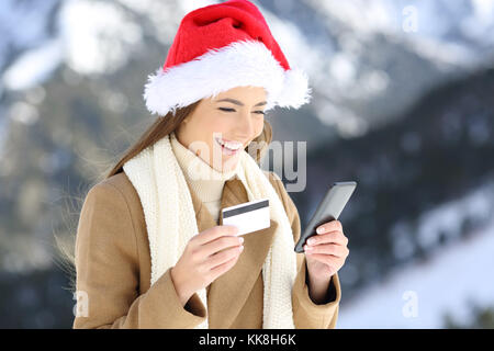 Portrait of a woman wearing santa claus hat on christmas paying on line with credit card and smart phone with a snowy mountain in the background Stock Photo