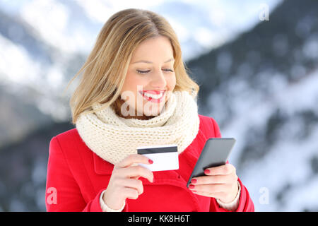 Portrait of a smiley woman paying with credit card and mobile phone in winter with a snowy mountain in the background Stock Photo