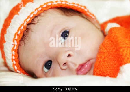 Pensive baby. Portrait of meditative newborn baby with beautiful blue eyes  in orange knitted costume on white fur blanket. Stock Photo