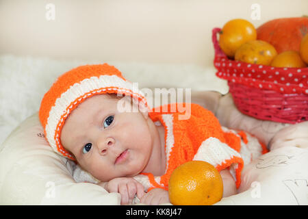 Cute few days newborn baby with funny curious face dressed in a knitted orange costume with oranges and pumpkin background on white blanket. Baby face Stock Photo