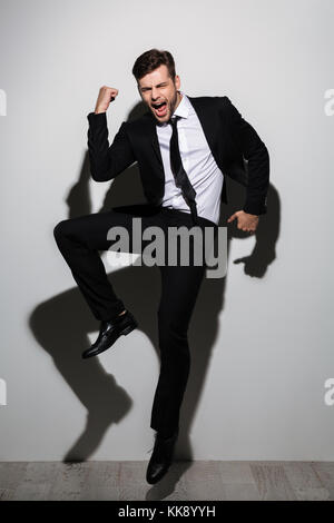 Full length photo of overjoyed handsome guy in black suit showing winner gesture while jumping over white background Stock Photo