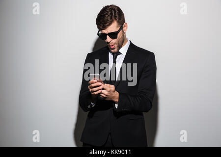 Close-up photo of amazed bearded man in black suit holding mobile phone, isolated on white background Stock Photo