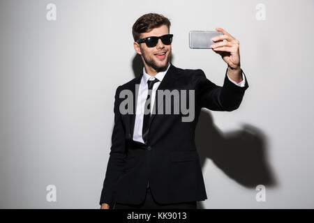 Cool bearded guy in black suit and suglasses taking selfie on mobile phone, isolated over white background Stock Photo