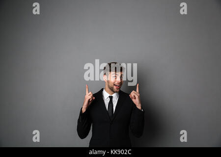 Happy attractive guy in black suit pointing with two fingers upward, isolated on gray background Stock Photo
