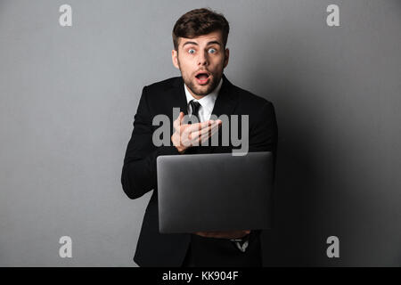 Amazed young guy in black suit holding laptop, looking at camera, isolated on gray background Stock Photo