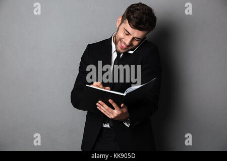 Happy bearded man in black suit taking notes while talking on smartphone, isolated on gray background Stock Photo
