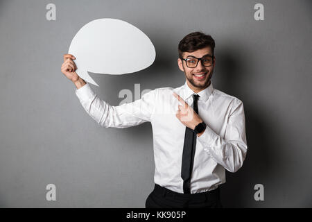 Happy handsome bearded man in white shirt pointing with finger on empty speech banner, isolated on gray background Stock Photo