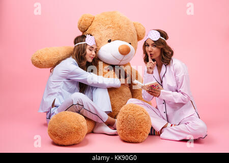 Two pretty girls dressed in pajamas sitting with a big teddy bear and reading a book isolated over pink Stock Photo