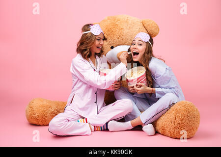 Two cheerful pretty girls dressed in pajamas sitting with a big teddy bear and eating popcorn isolated over pink background Stock Photo