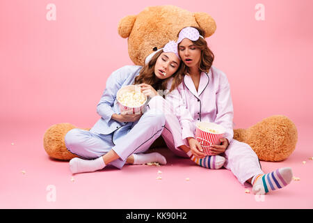 Two pretty girls dressed in pajamas sitting with a big teddy bear and sleeping with popcorn isolated over pink background Stock Photo