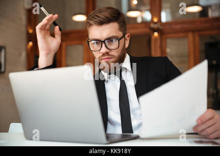 Young concentrated bearded businessman in formalwear looking at laptop screen holding papers while sitting at workplace Stock Photo