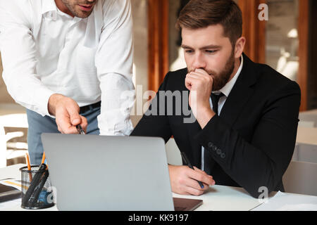 Close-up photo of two young man sitting in office and discussing business using laptop Stock Photo