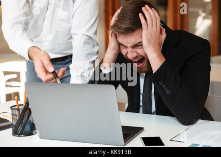 Young angry bearded businessman in formalwear holding his head, looking at computer while his partner pointing with pen on laptop screen in office Stock Photo