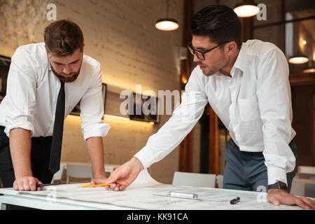 Two young serious businessmen working on a business plan while standing over desk with graph indoors Stock Photo