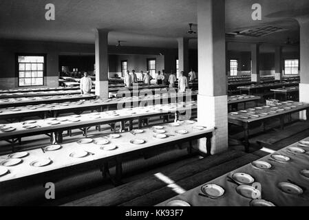 Black and white photograph of a large dinning hall, empty except for a group of staff, place settings consist of worn enameled plates, a fork and a knife, by Edwin Levick, Ellis Island, New York, 1907. From the New York Public Library. Stock Photo