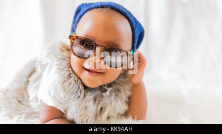 Super cute cool baby. Hipster baby in fur vest and sunglasses lies on a white bed in a room with curtains. Baby smiles while putting on sunglasses. Stock Photo