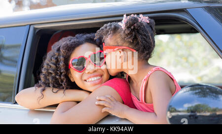 Summer fun! Happy girls or sisters with heart shaped sunglasses in car window. Younger girl kisses older girl on cheek in celebration of love, summer  Stock Photo